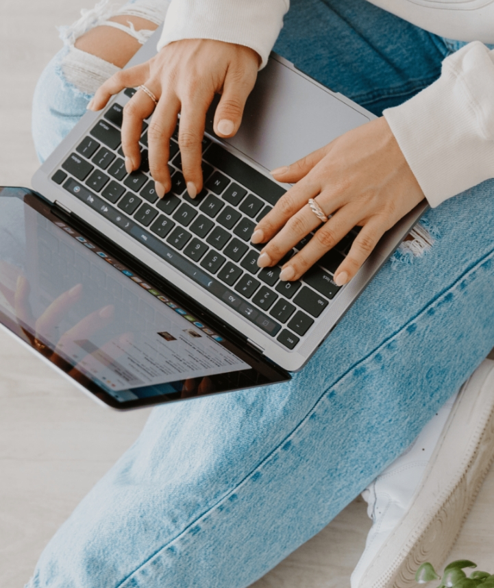 An upwards angle of a woman typing on a MacBook while sitting down. Her laptop is rested on her lap and she is wearing blue jeans.